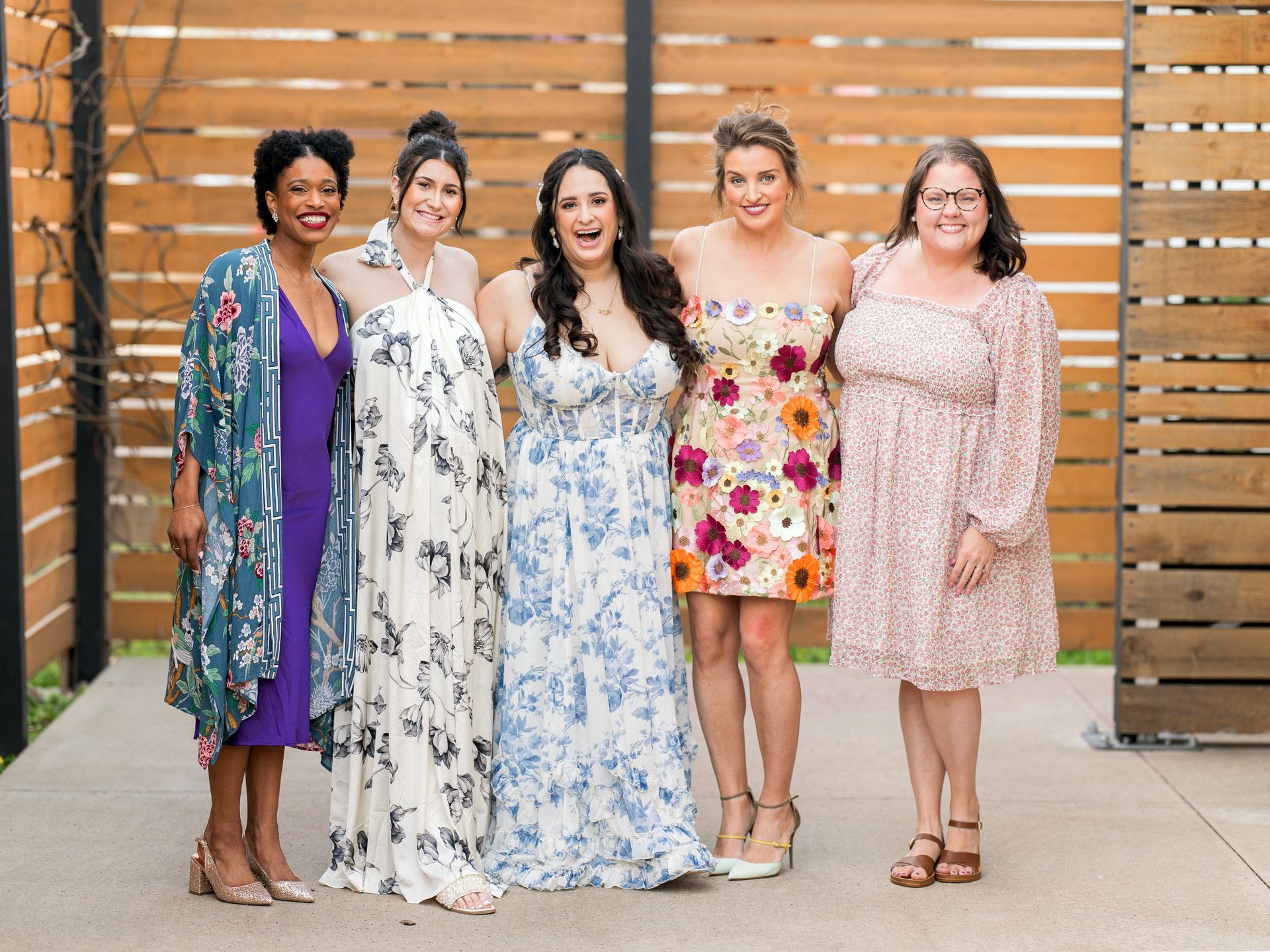 Five women in floral dresses pose outdoors in front of a wood fence, smiling.