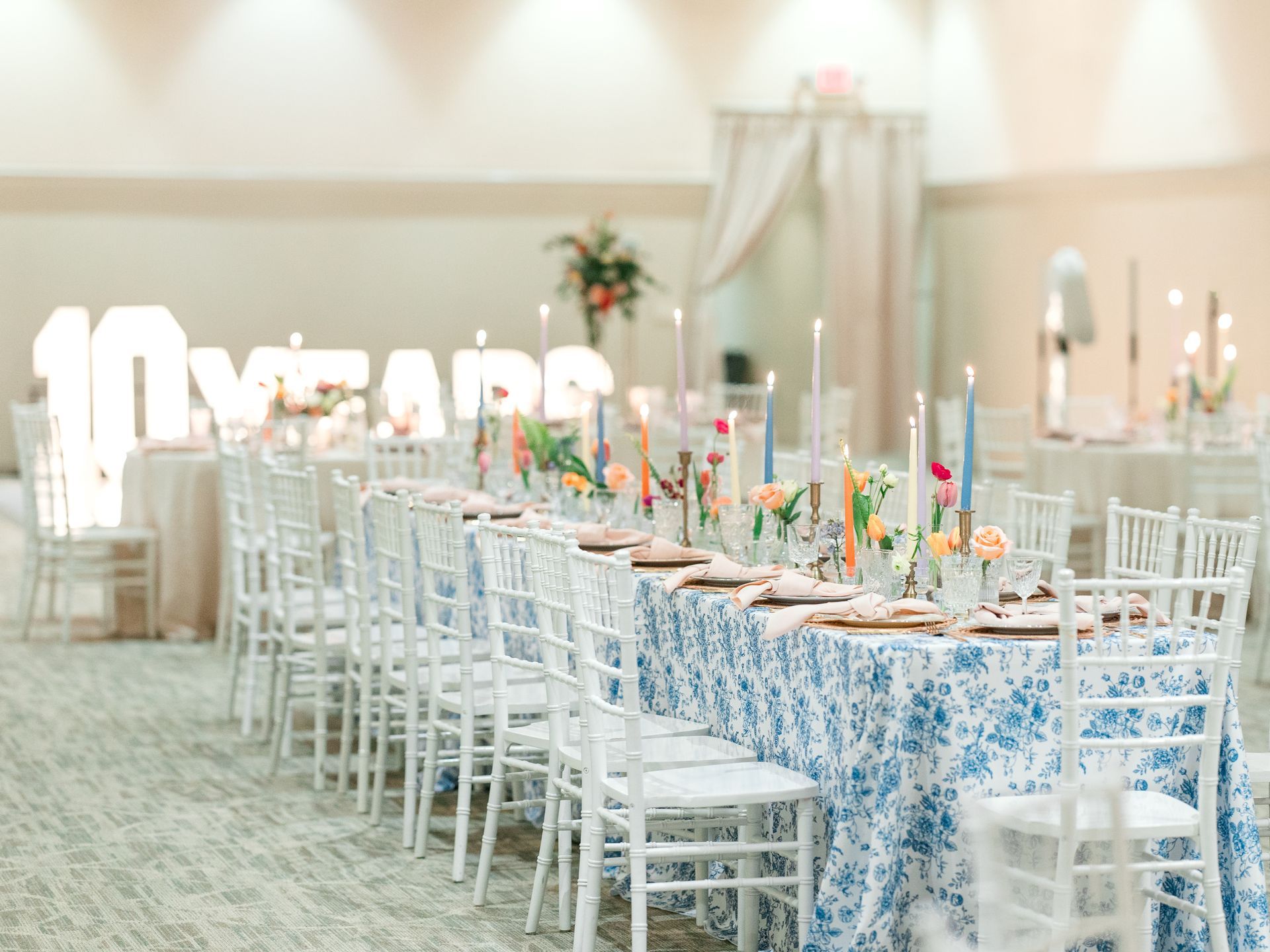 Long table set for celebration, floral centerpiece, blue patterned tablecloth, lit candles, white chairs, and large “10 YEARS” sign.