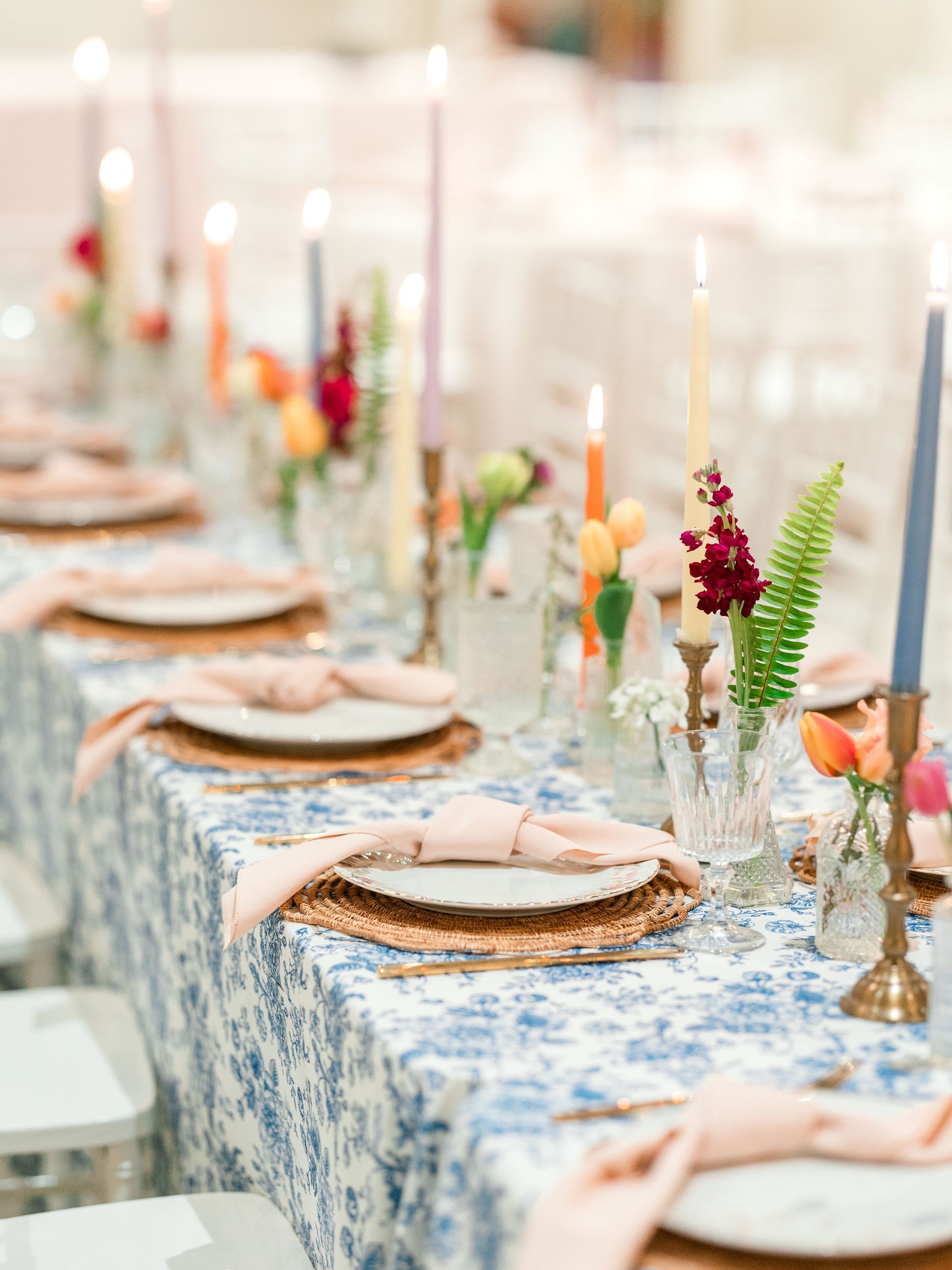 Table setting with blue floral tablecloth, gold chargers, pink napkins, and colorful candles.