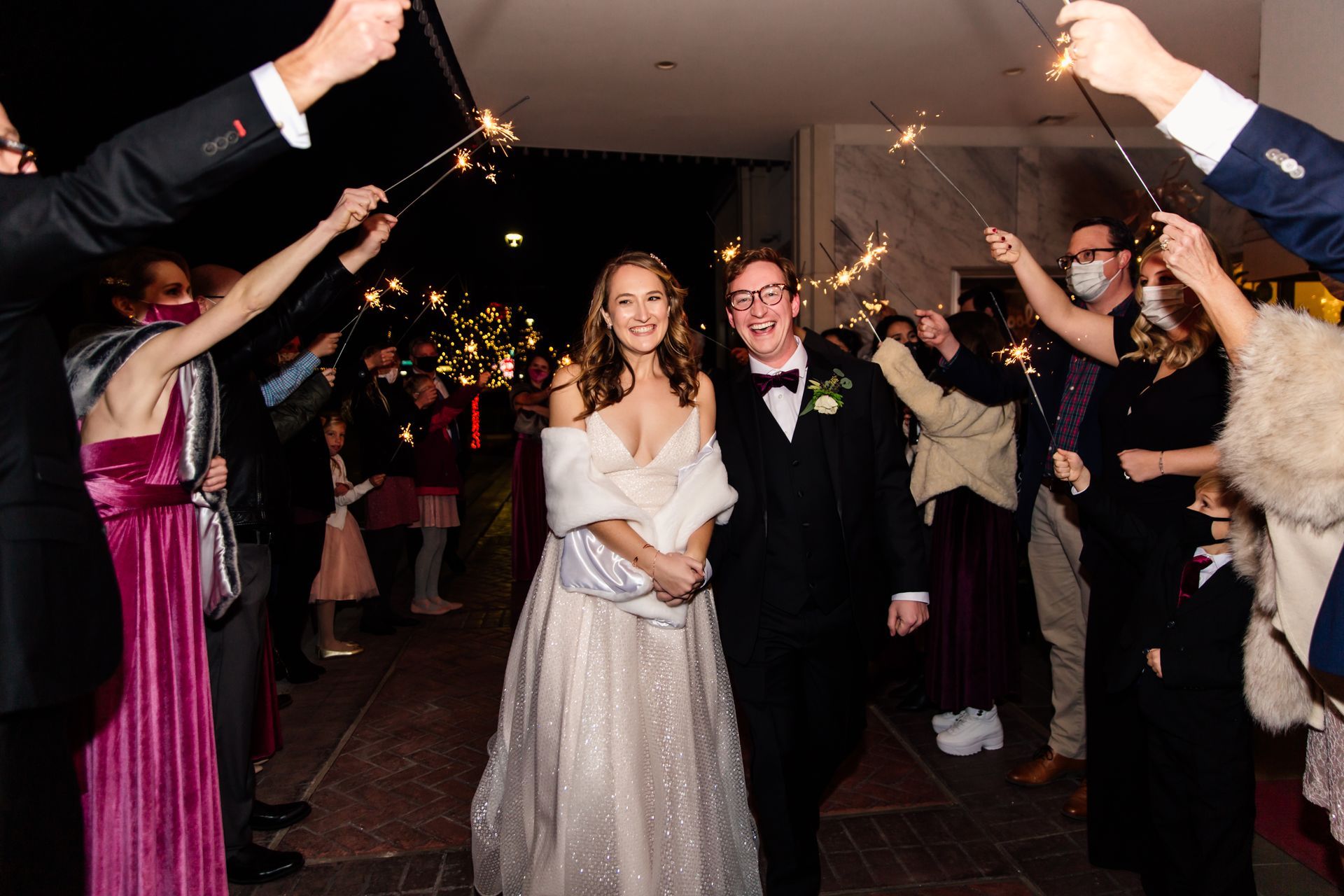 Bride and groom walk through sparklers held by guests after a wedding. Evening, joyful.