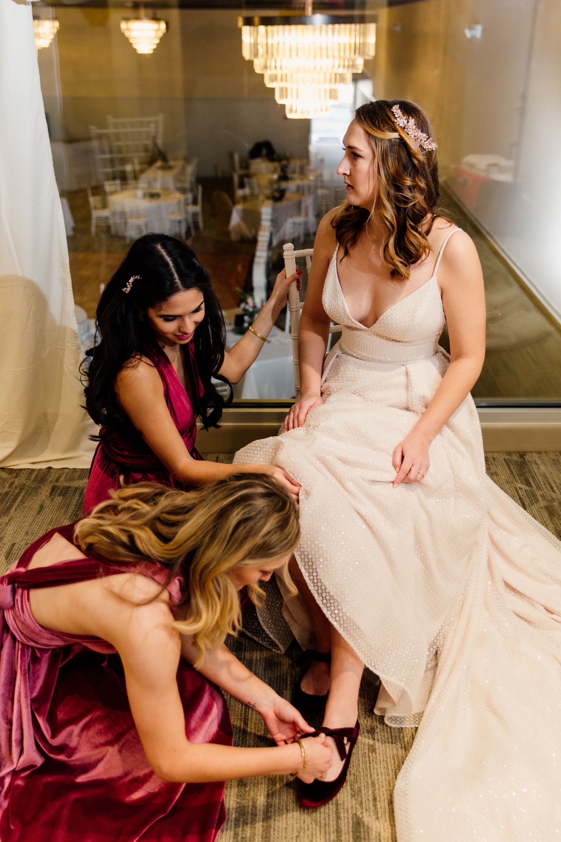 Bride having shoes tied by bridesmaids; indoor setting with large window.