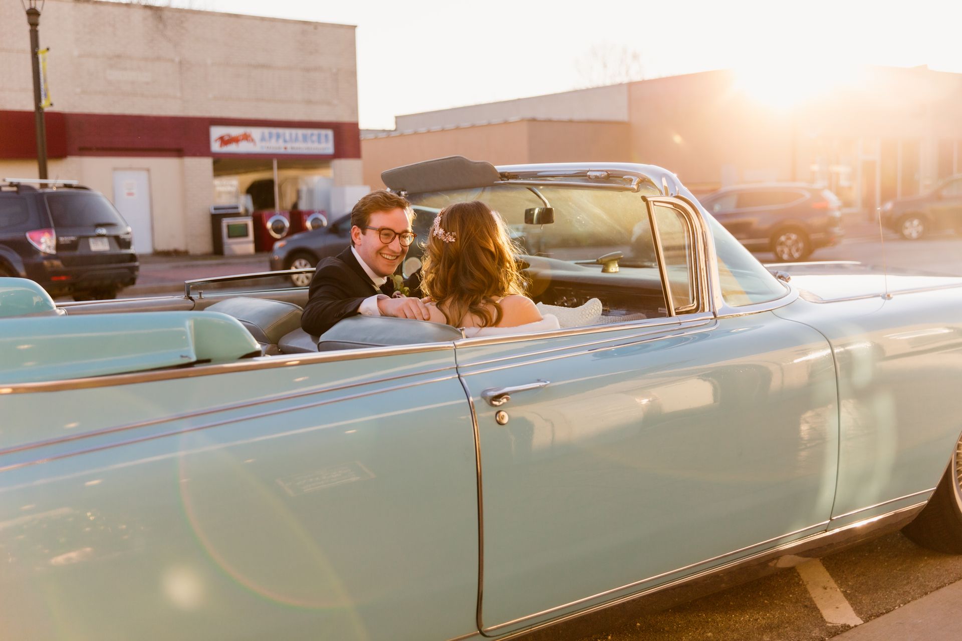 Couple in formal attire smiles in a vintage turquoise convertible, setting sun in background.