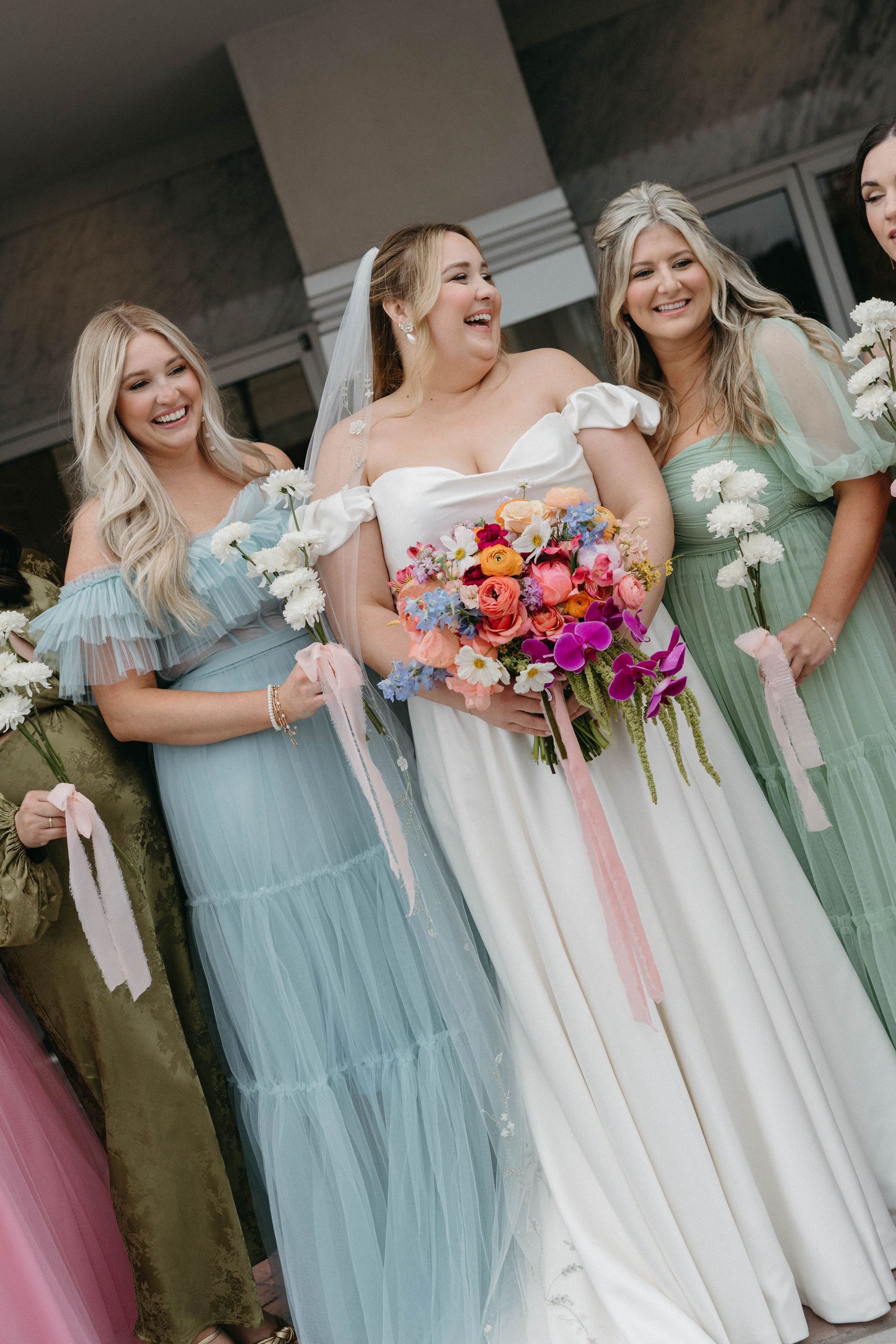 Bride smiles with bridesmaids, holding flowers and ribbons. Dresses are blue, green, and pink.