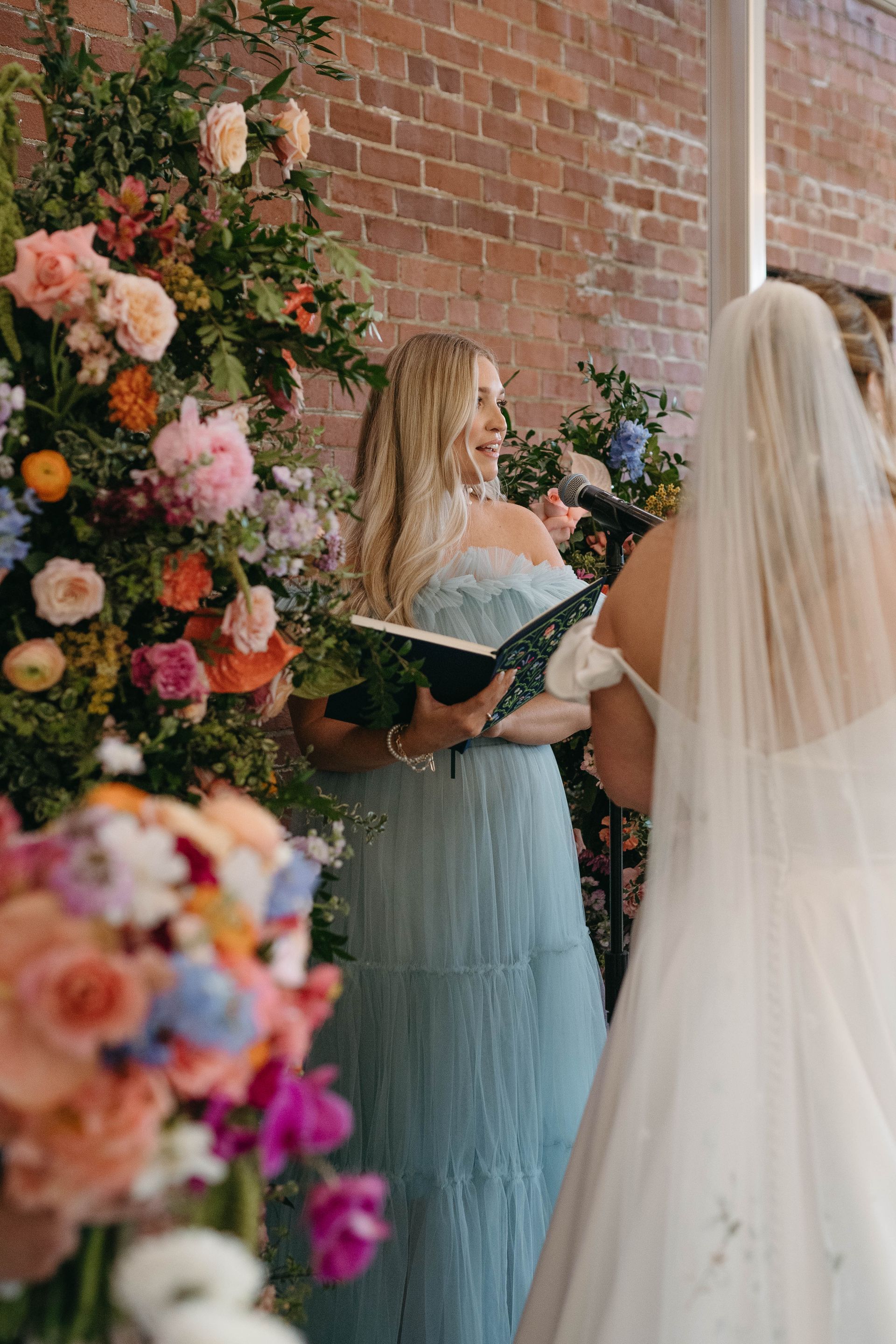 Woman in blue dress reads at a wedding ceremony; bride in white gown stands nearby.