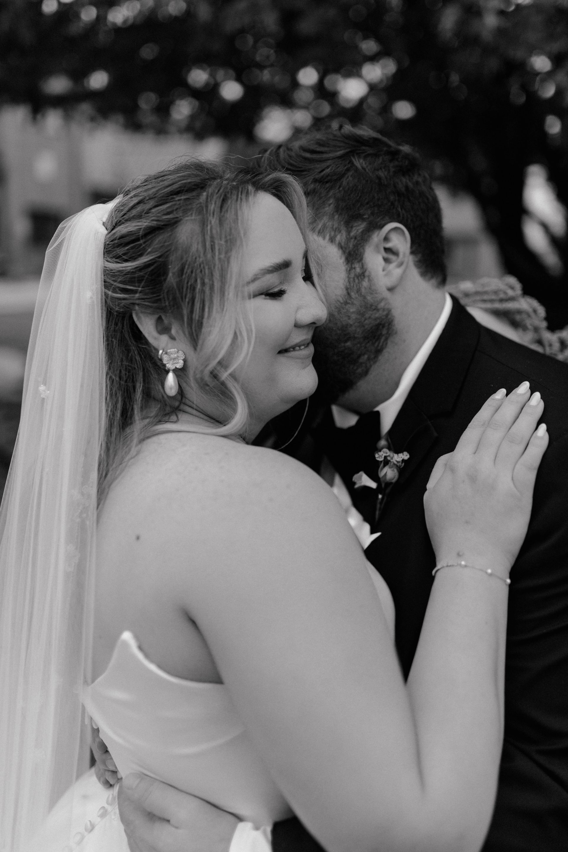 Bride and groom embrace outdoors; the bride wears a veil and the groom a suit. Black and white photo.