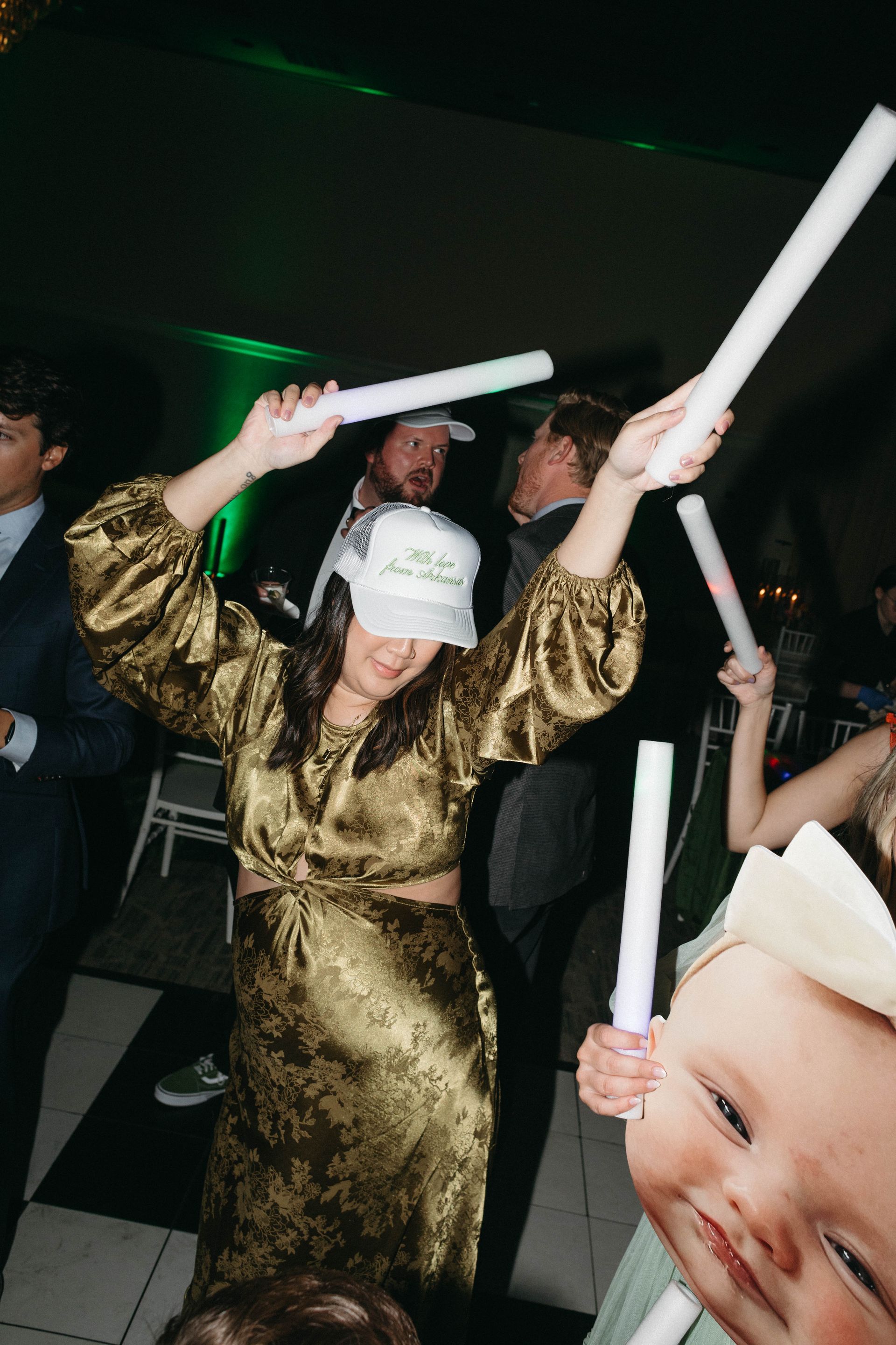 Woman in gold dress dancing, holding glow sticks at an event, wearing a hat with text. Others also have glow sticks.