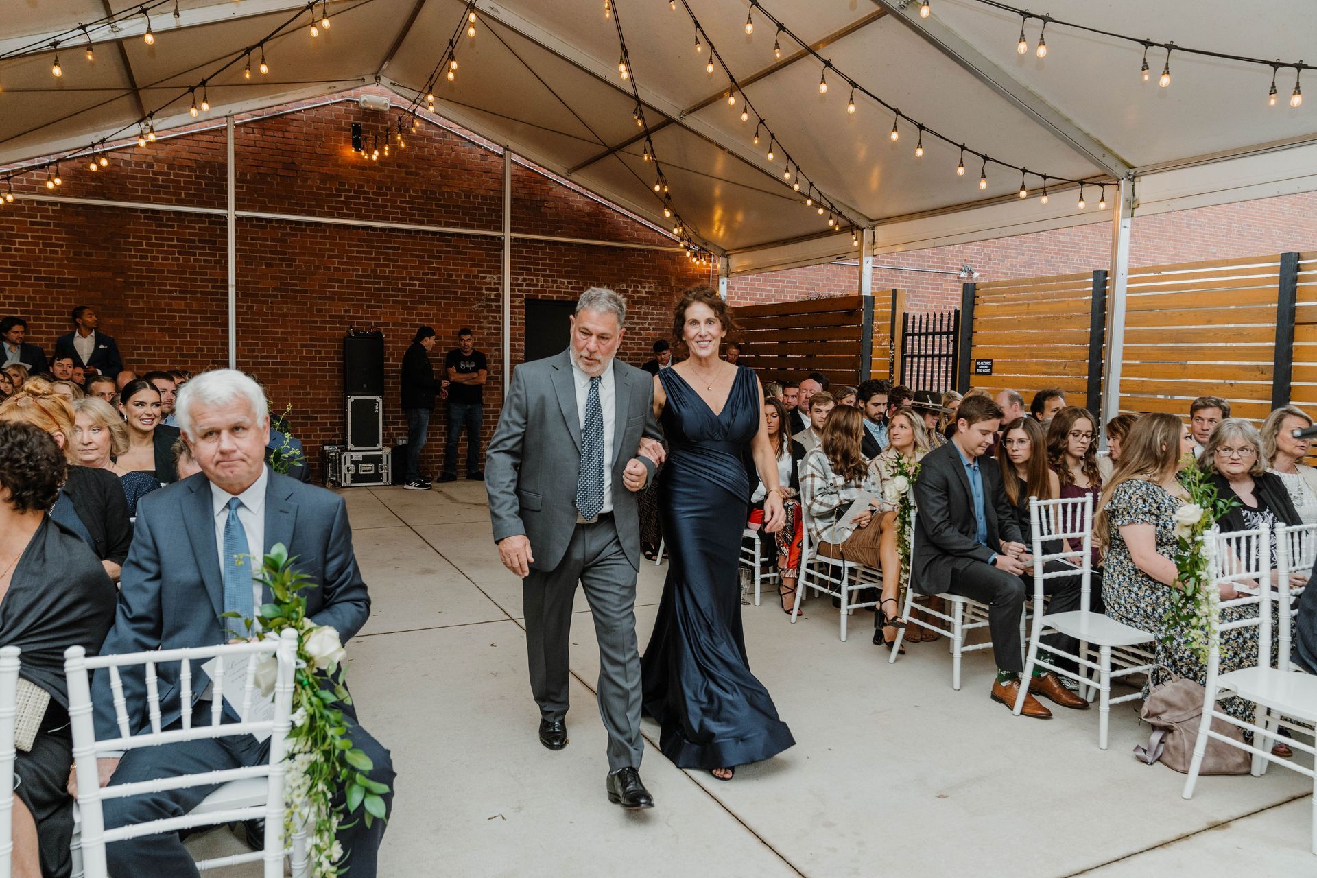 A woman in a navy dress is escorted down an aisle by a man at an outdoor wedding.