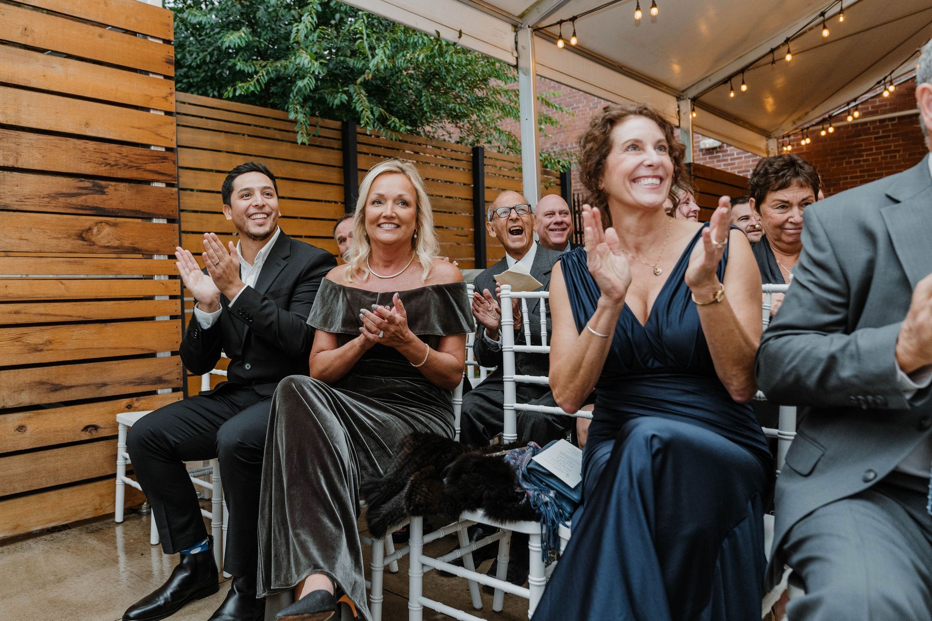 People clapping at a formal outdoor event; smiles, dark blue and gray formal wear.