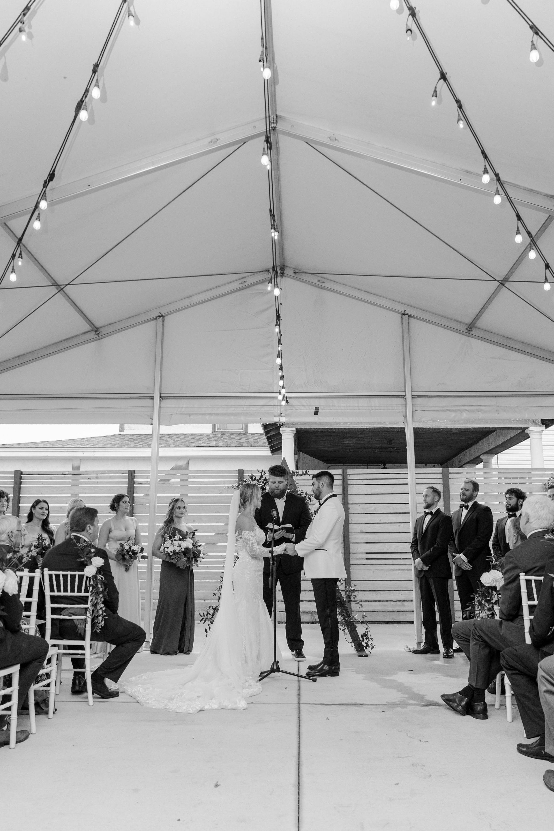 Wedding ceremony under a tent with string lights. Couple kissing, officiant, wedding party, and guests seated.