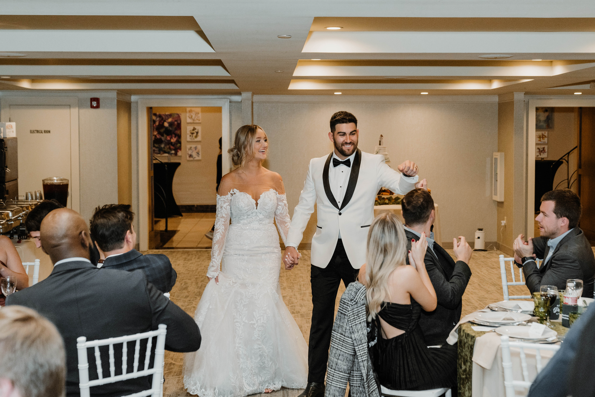 Newlyweds enter reception, holding hands, smiling, in formal wear. Guests applaud at tables in a banquet hall.