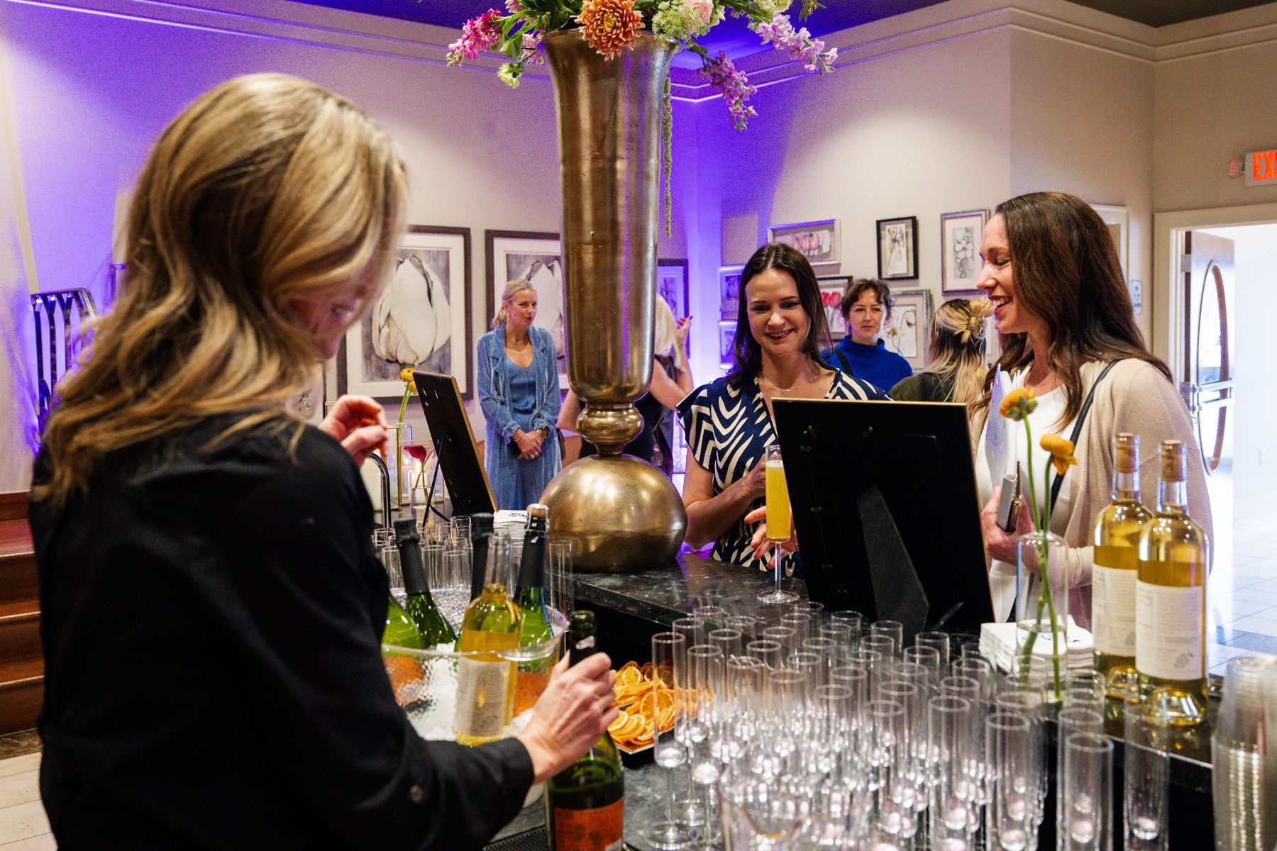 People at a bar, pouring drinks. Woman behind bar, two customers. Elegant setting, flowers, bottles, glasses.