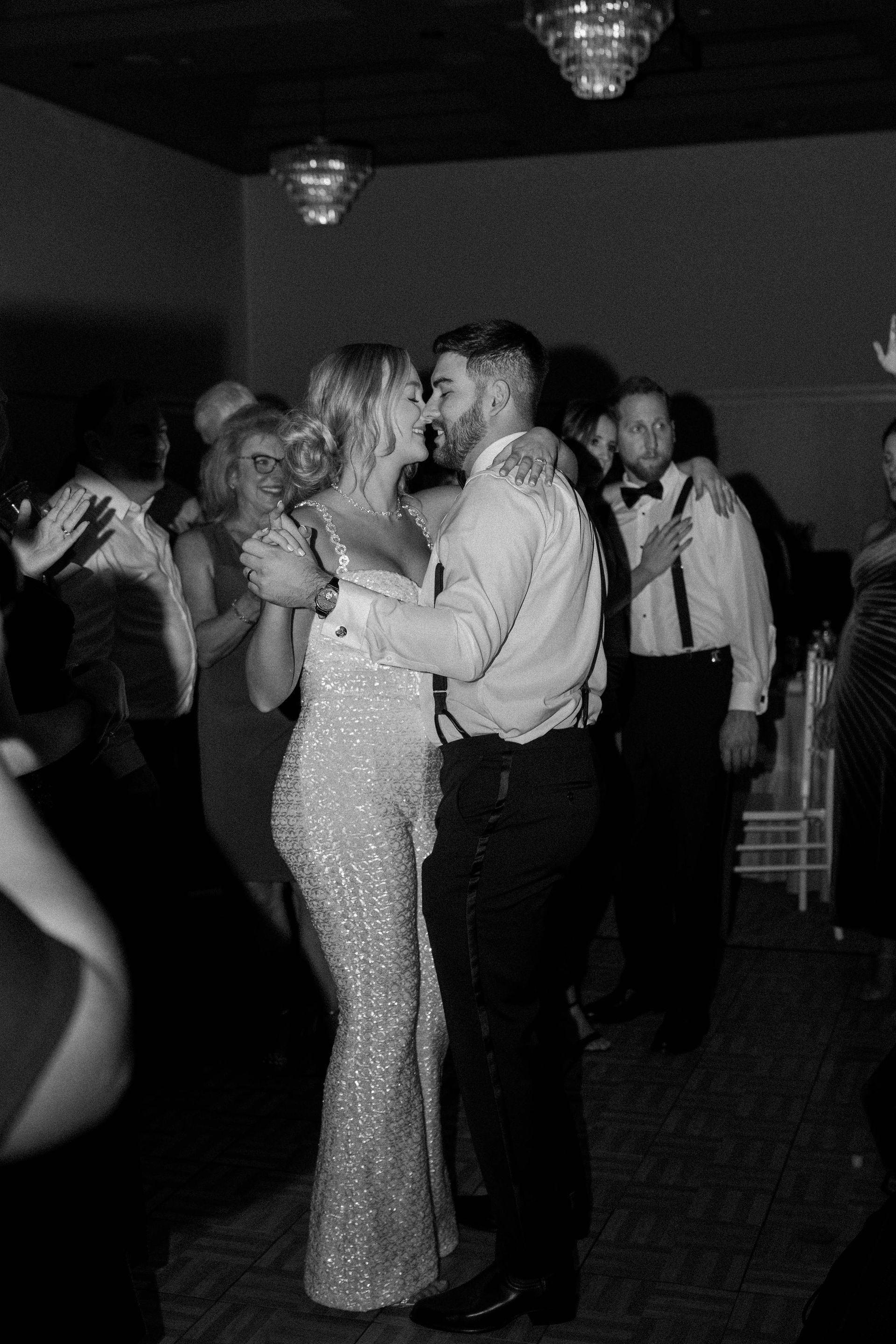 Couple dancing at a formal event; woman in a sequined gown, man in suspenders, soft lighting.