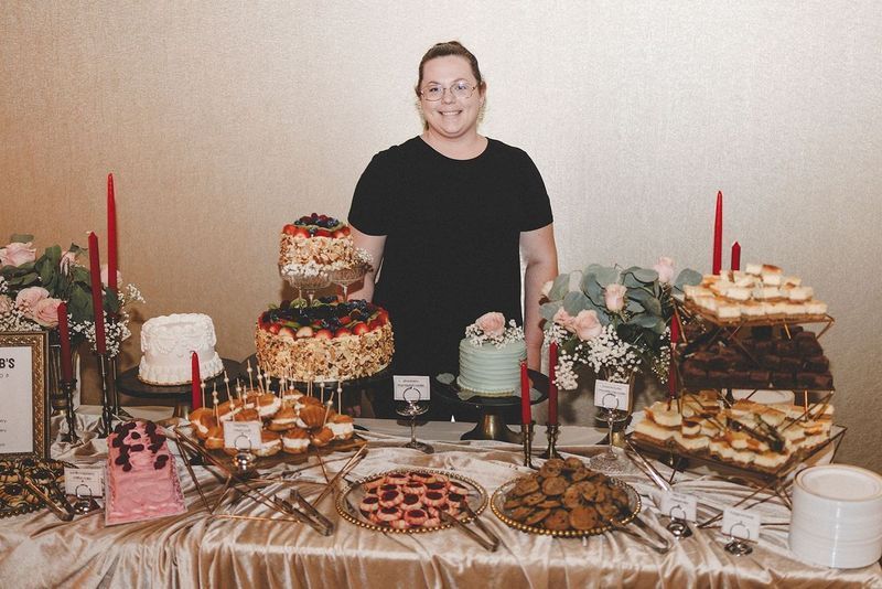 Person stands behind a dessert table with multiple cakes, pies, and pastries.