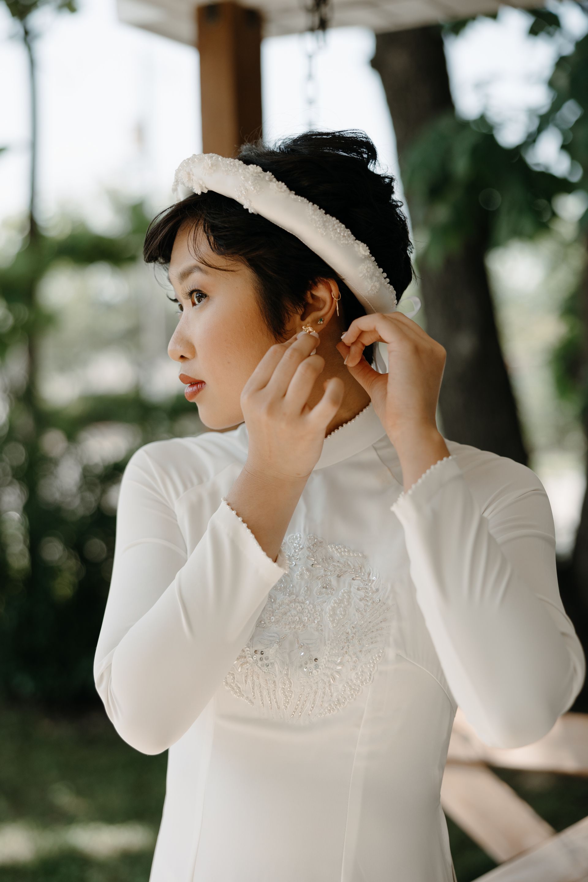 Woman adjusting an earring, wearing a white ao dai and headpiece outdoors.