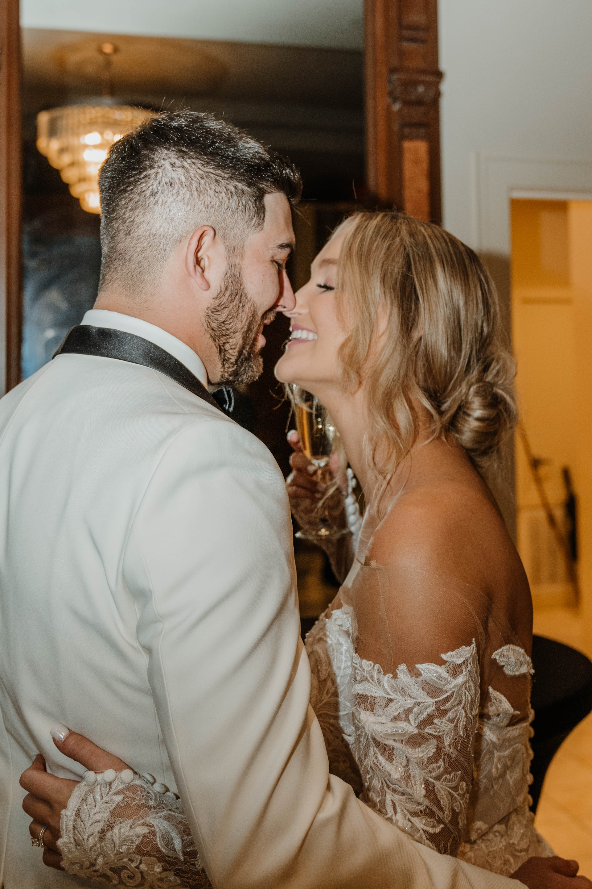 Bride and groom embrace, smiling, holding champagne. Bride's dress has lace sleeves. Groom wears a white suit.