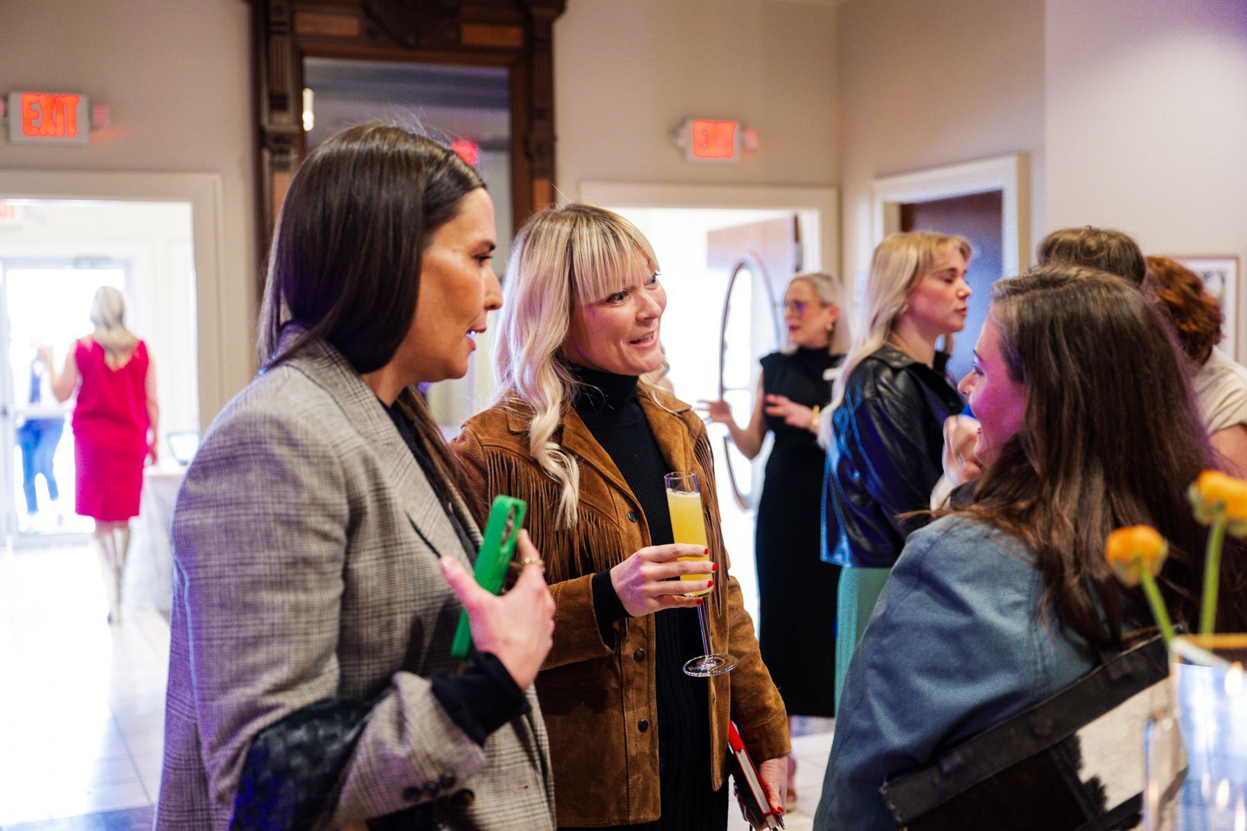 People socializing at an event. Women talking, one holding a drink. Brightly lit room.