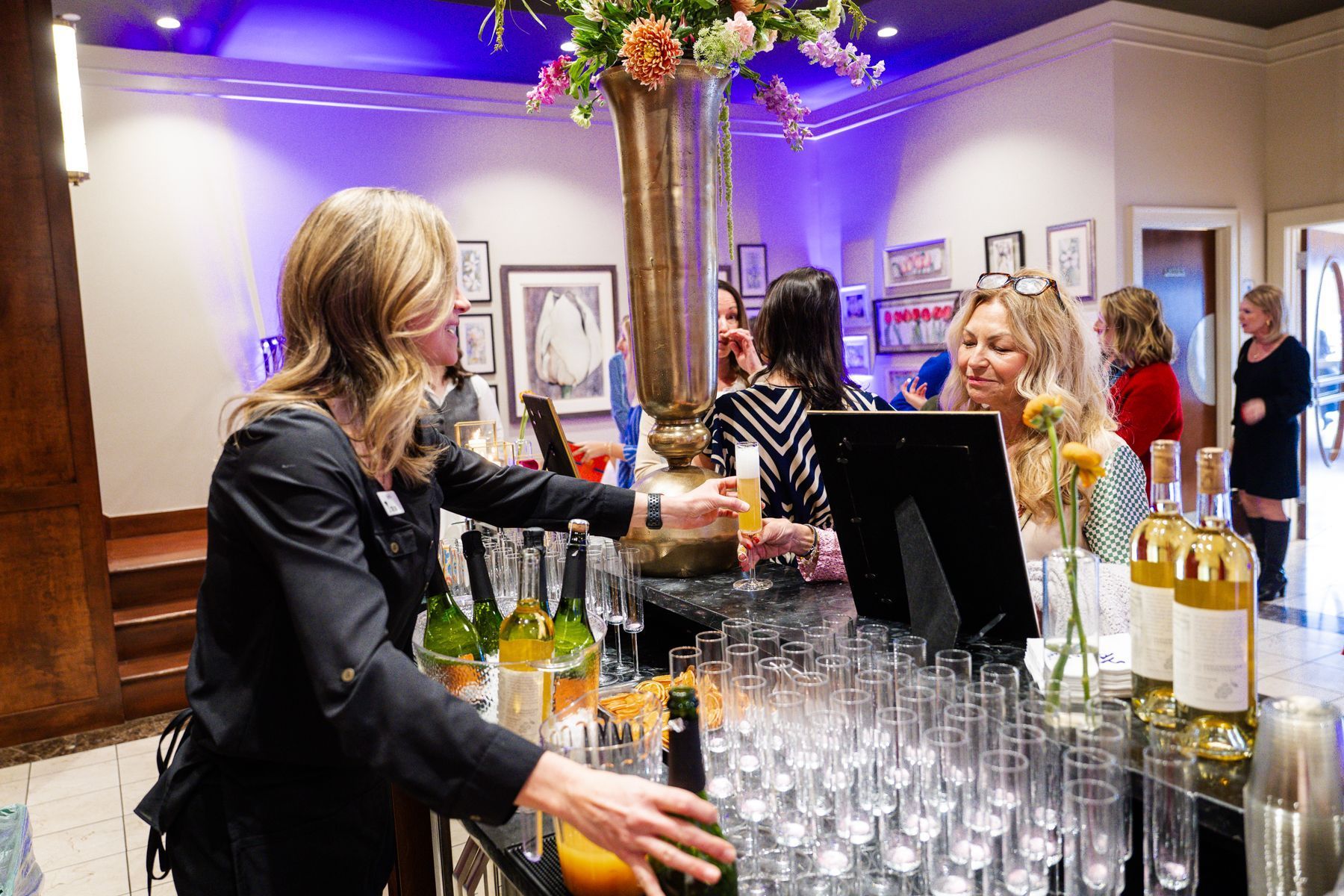 Woman serving drinks at a bar, with several people gathered. The setting is indoors, with floral arrangements.