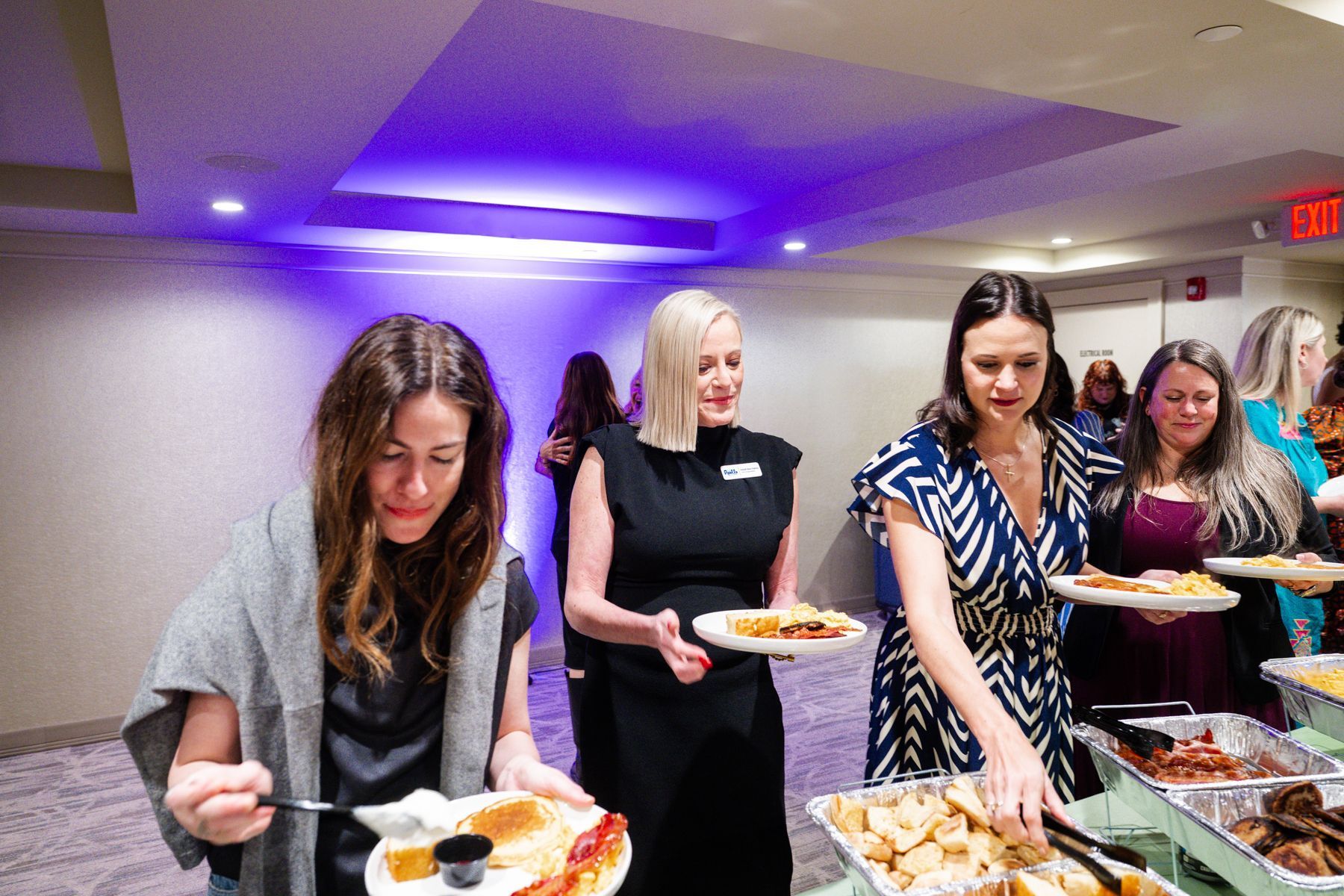People serving themselves food from a buffet at an event, inside a room with purple lighting.