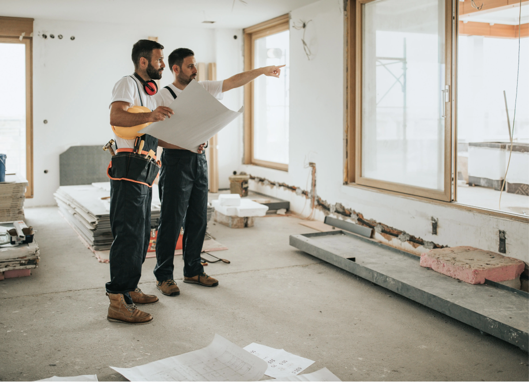 Two construction workers reviewing blueprints in a room under renovation. One points, the other holds plans.