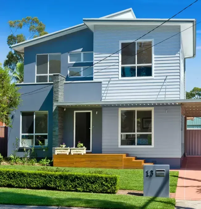 Two-story modern house with grey and blue exterior. Wooden deck, green lawn, and street address