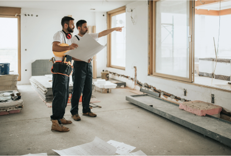 Two workers reviewing a blueprint in a room under renovation, with tools and materials scattered around.