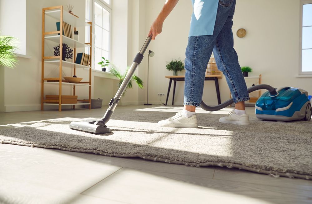 Person vacuuming a gray carpet in a bright, sunny room.