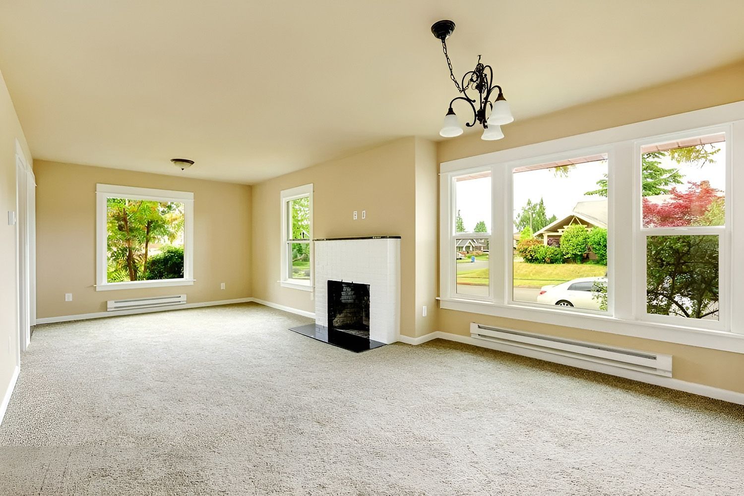 Bright empty living room with beige walls, fireplace, carpet, and large windows overlooking greenery