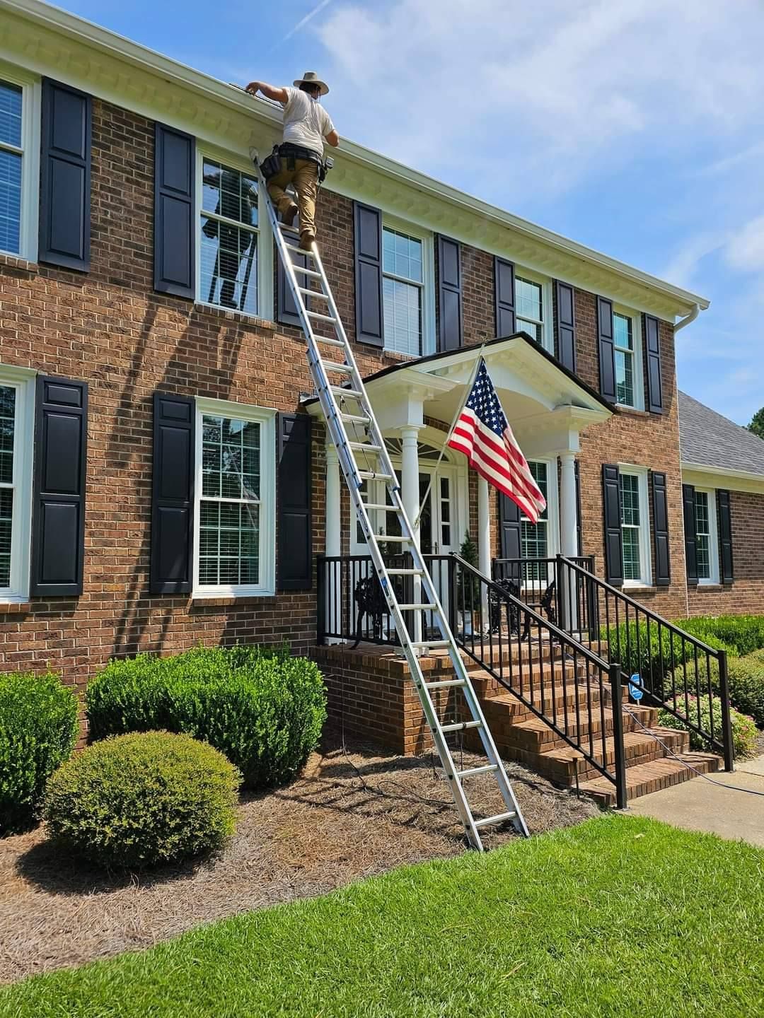 A man is standing on a ladder cleaning the windows of a brick house.