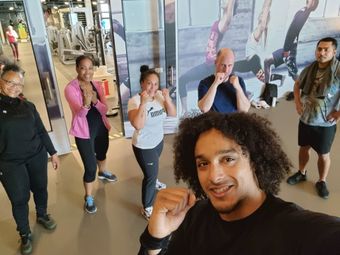 Groep in de sportschool, sommigen met gebalde vuisten, lachend. Man op de voorgrond maakt een selfie. Fitnessapparatuur en muurschilderingen.