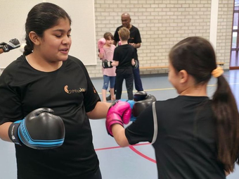Twee kinderen boksen, een van hen deelt een stoot uit. Training in een sportschool; instructeur op de achtergrond.