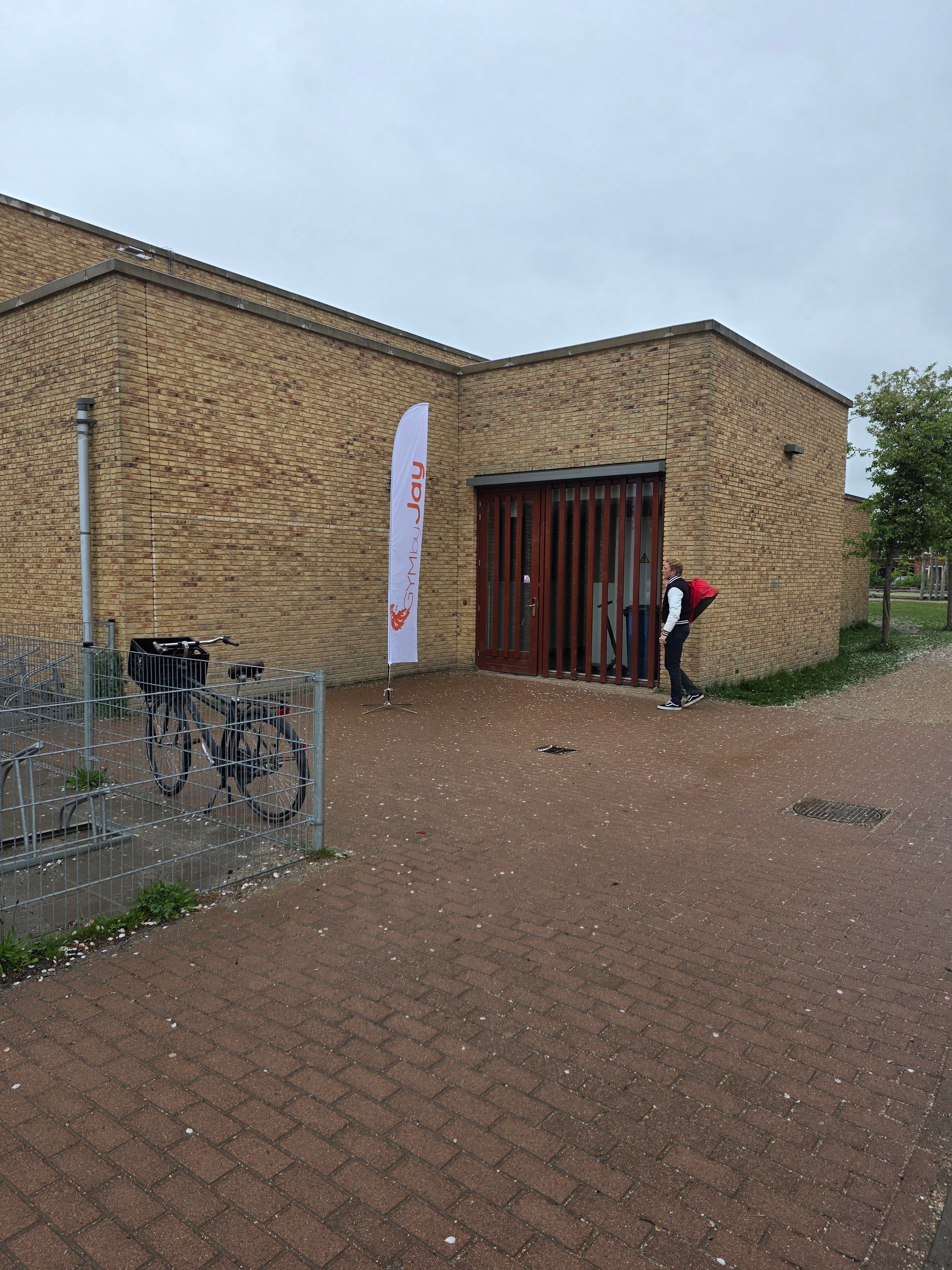 Bakstenen gebouw met houten deur, spandoek en persoon met rugzak. Fietsen en kleine witte voorwerpen op de grond.