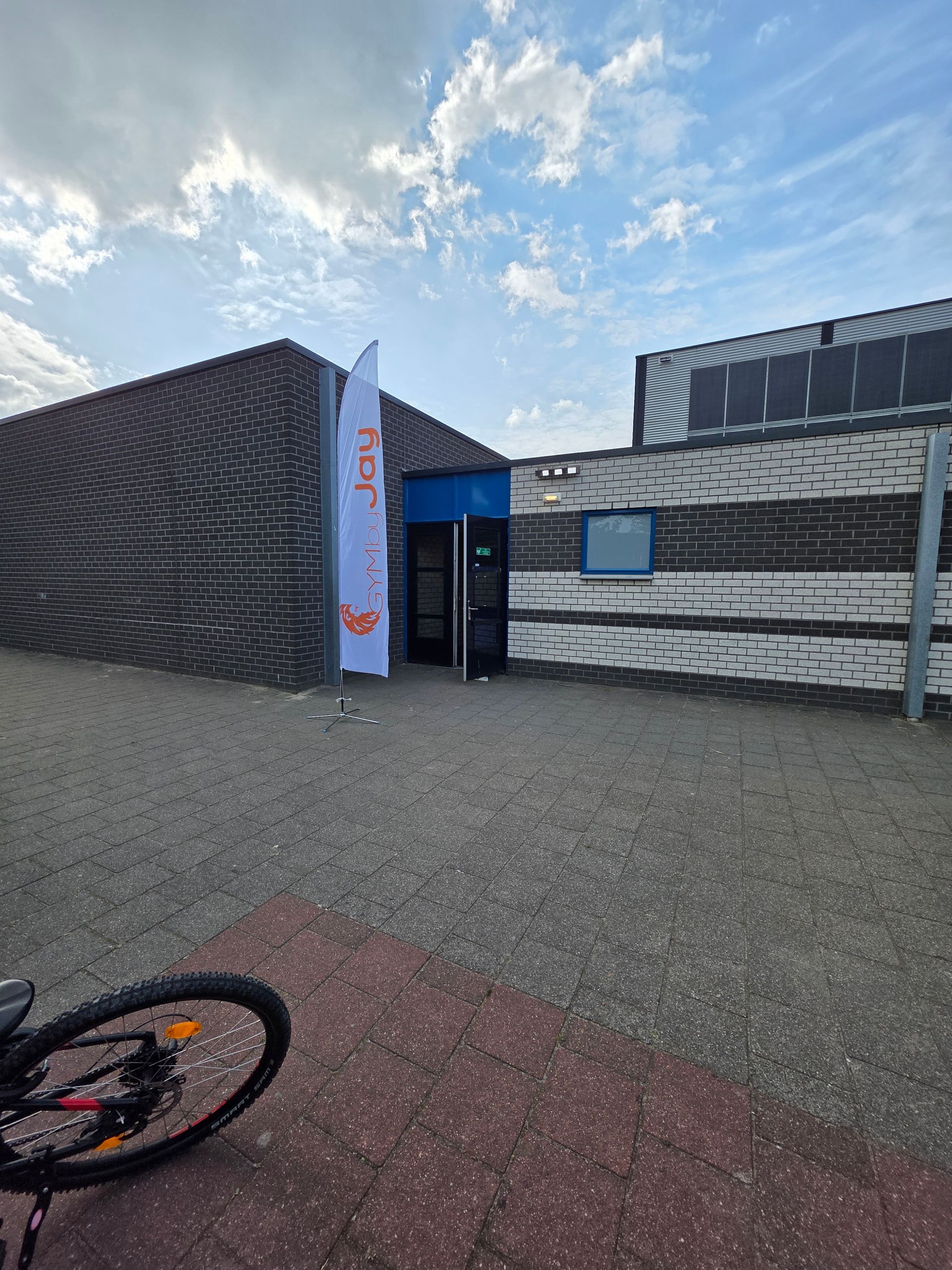 Ingang van een gebouw met een vlag; deur open. Grindpad, bakstenen en houten muren. Een fietswiel op de voorgrond.