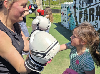 Vrouw met zwarte bokshandschoenen lacht naar kind tijdens een buitensportevenement met een beschilderde muur op de achtergrond.