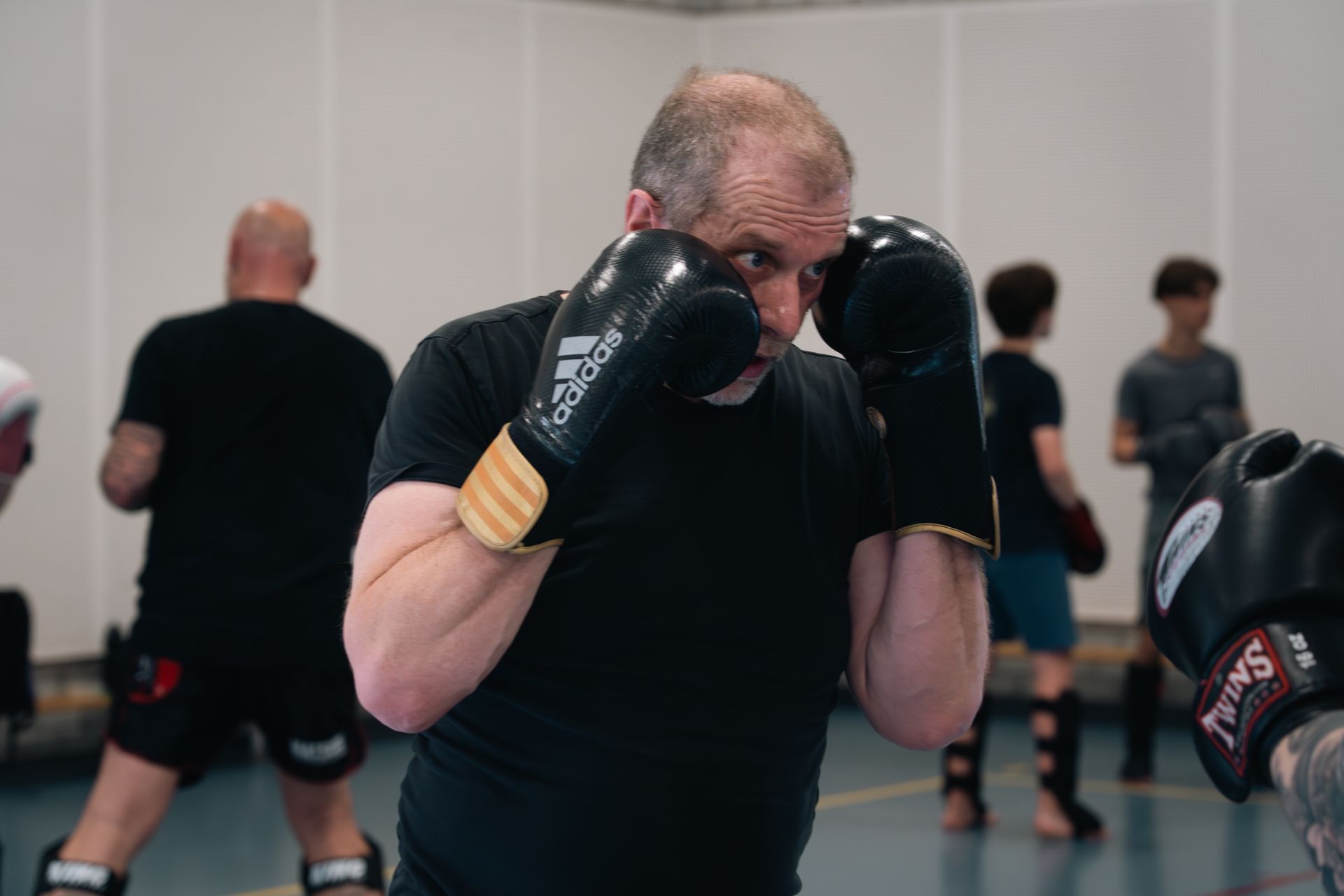 Boxer in black gloves and shirt sparring in a gym, with others training blurred in the background