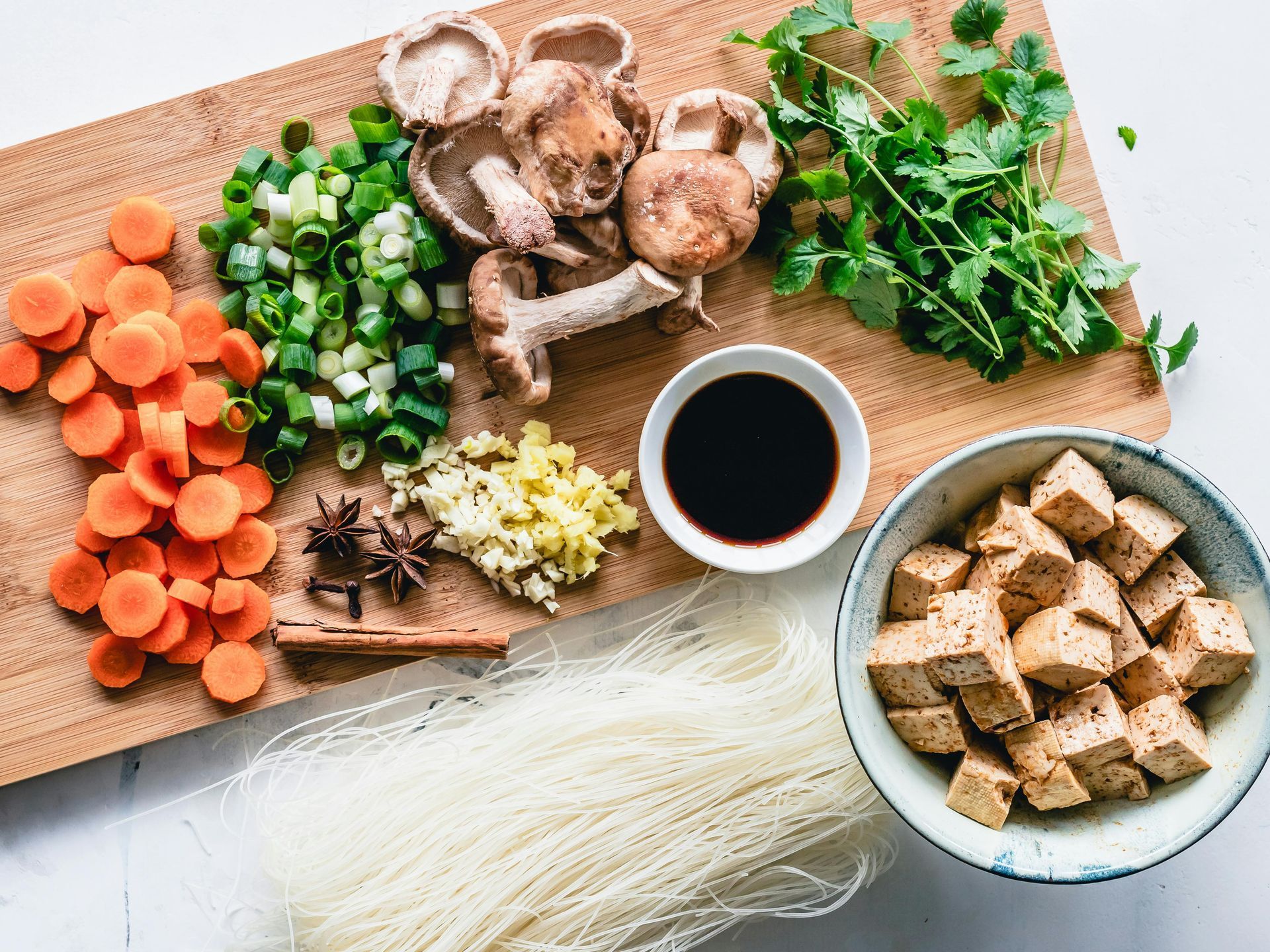 A wooden cutting board topped with noodles , carrots , mushrooms , and other vegetables.