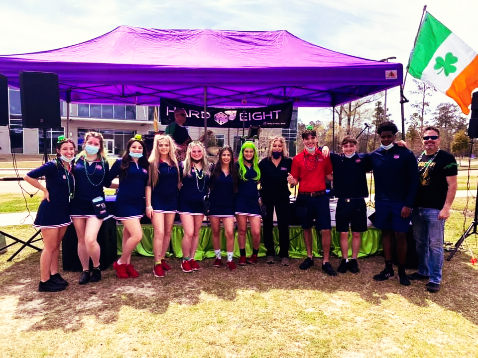 Group of people pose under purple tent with Irish flag, celebrating St. Patrick's Day outdoors.