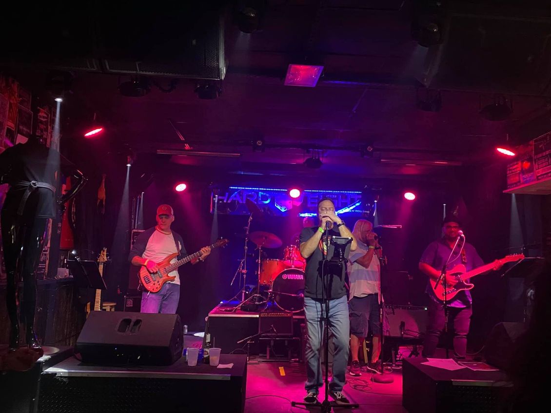 Band performing on stage under red and purple lights. Musicians with instruments, some singing into microphones.