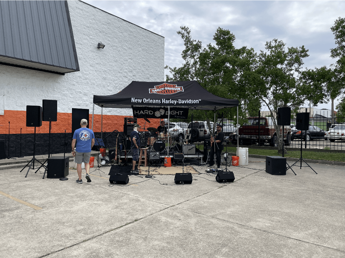 Band playing under a canopy at an outdoor event. Speakers and equipment are set up; a person stands nearby.
