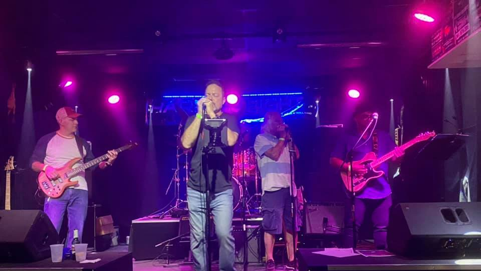 Band on stage performing under purple and blue stage lights. Man plays harmonica.