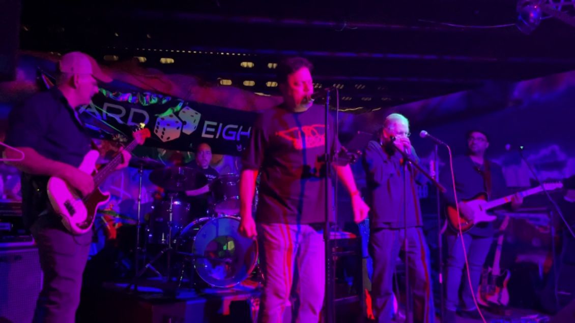 Band performing live on stage at a club with blue and purple lighting; vocalist with arms raised.