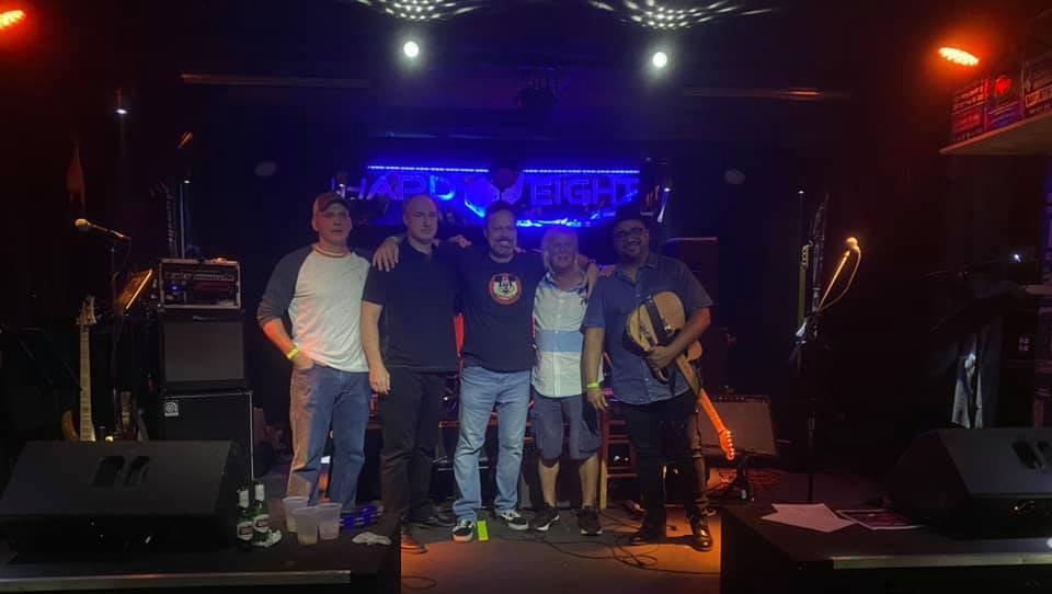 Band of five men posing on a stage, stage lights illuminating. Instruments in background.