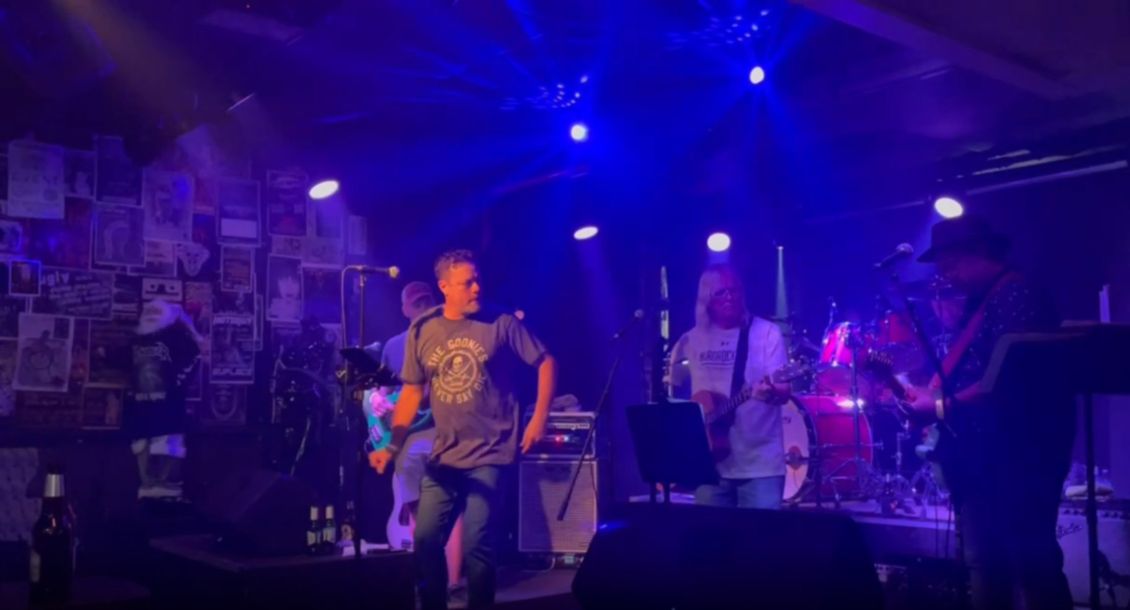 Band performing live on stage under blue stage lights. Musicians playing instruments, singer in front.