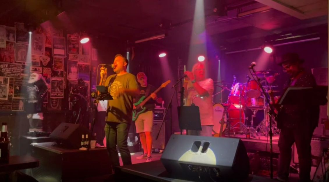 Band performing on stage under red and white lights. Musicians with instruments; audience in a dark setting.