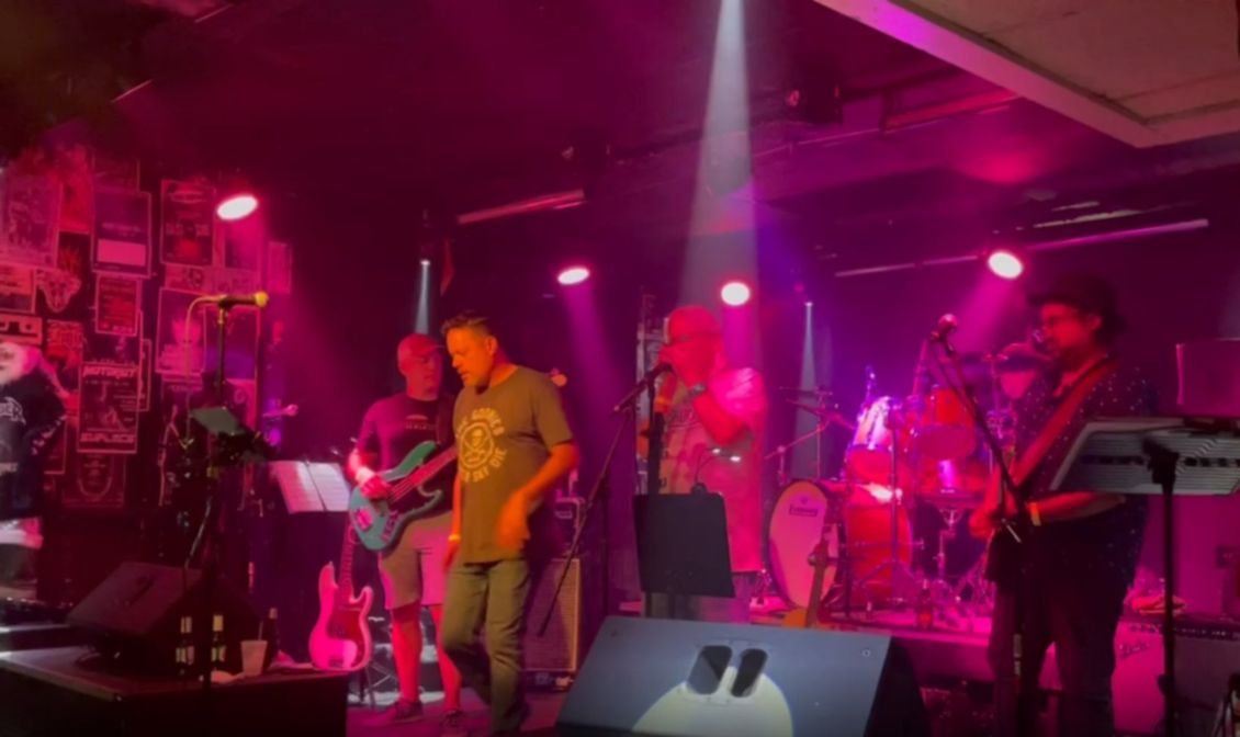 Band performing on a stage lit with pink and white lights. Musicians with instruments and microphones are visible.