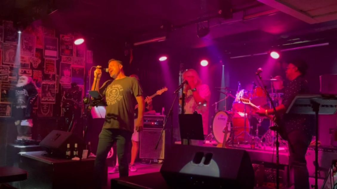Band performing live on stage with purple lighting. Singer at the front with a microphone, drummer, guitarist and keyboard player visible.