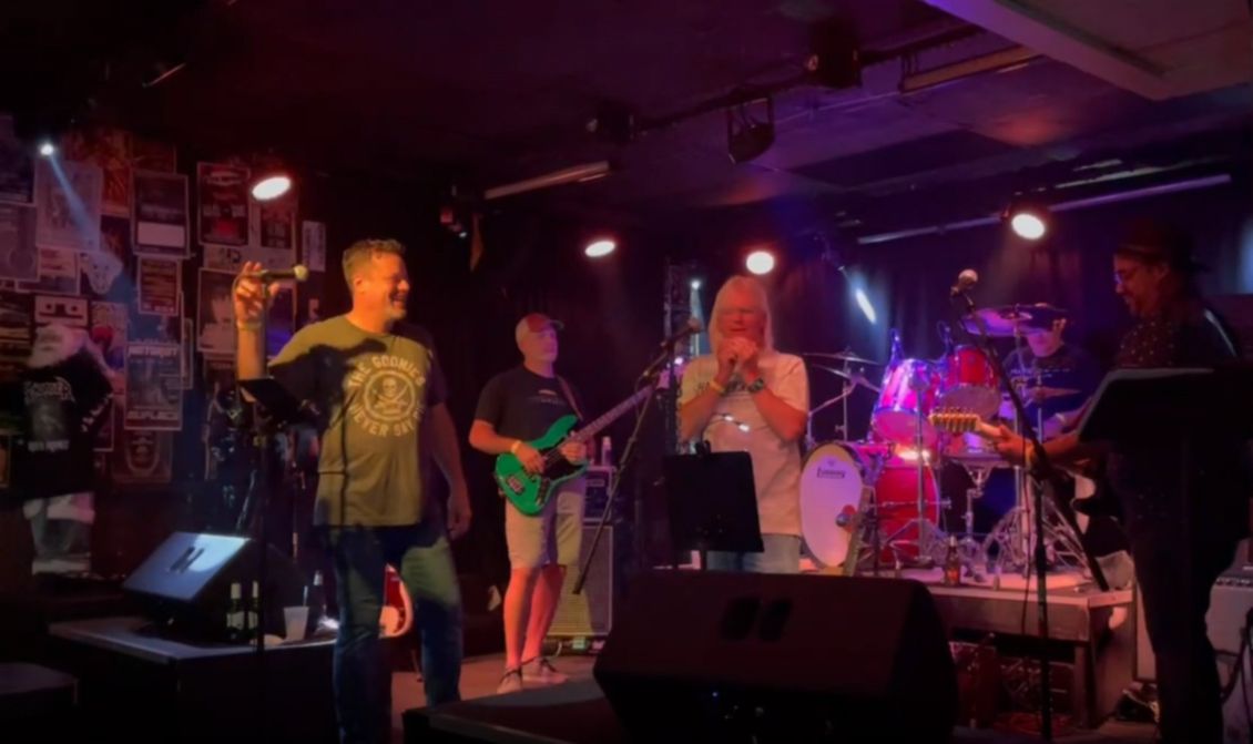 Band performing live in a dimly lit venue: singer with microphone, bassist, trumpeter, drummer, guitarist.
