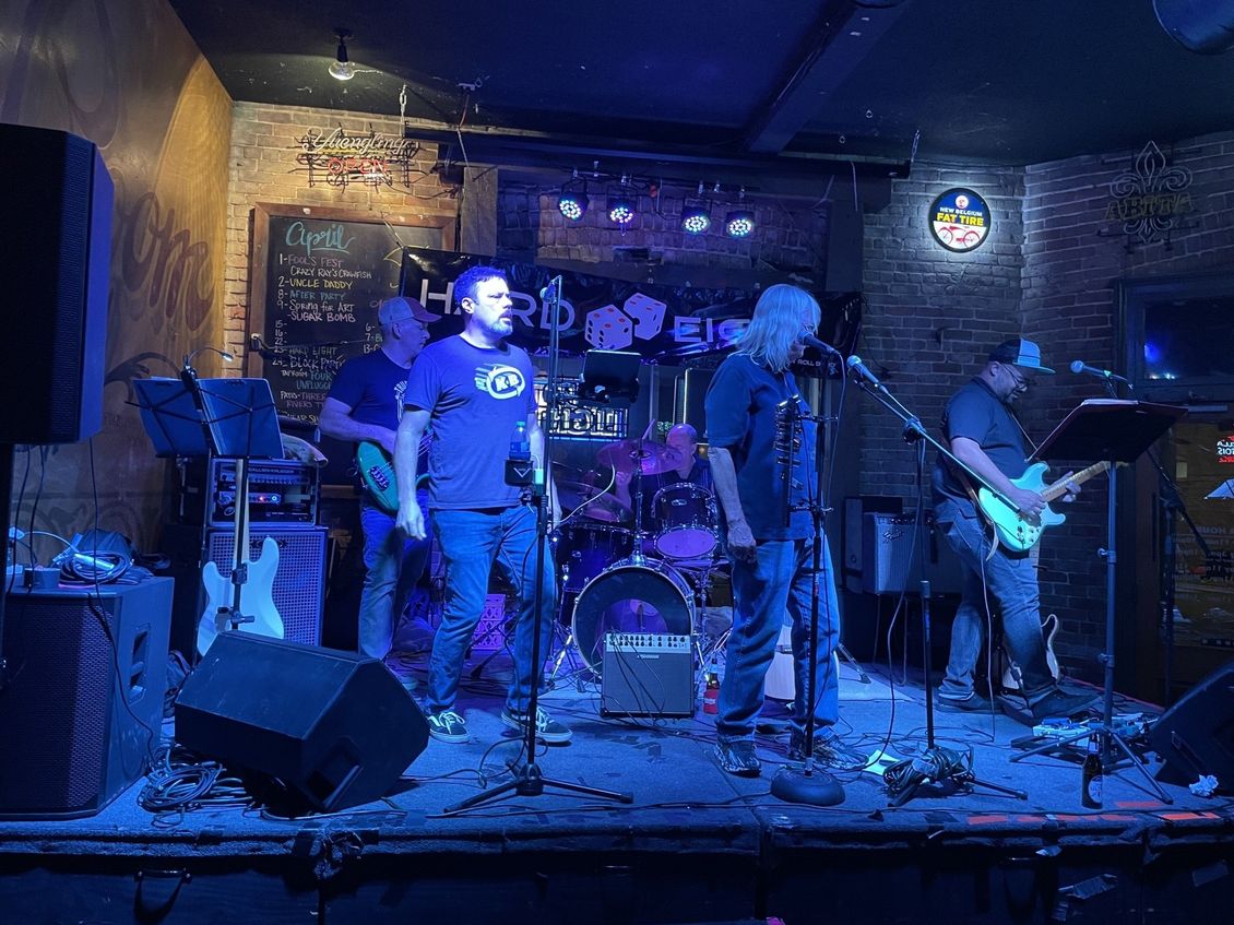 Band performing on stage in a dimly lit bar, under blue stage lights. Musicians playing instruments, singing into mics.