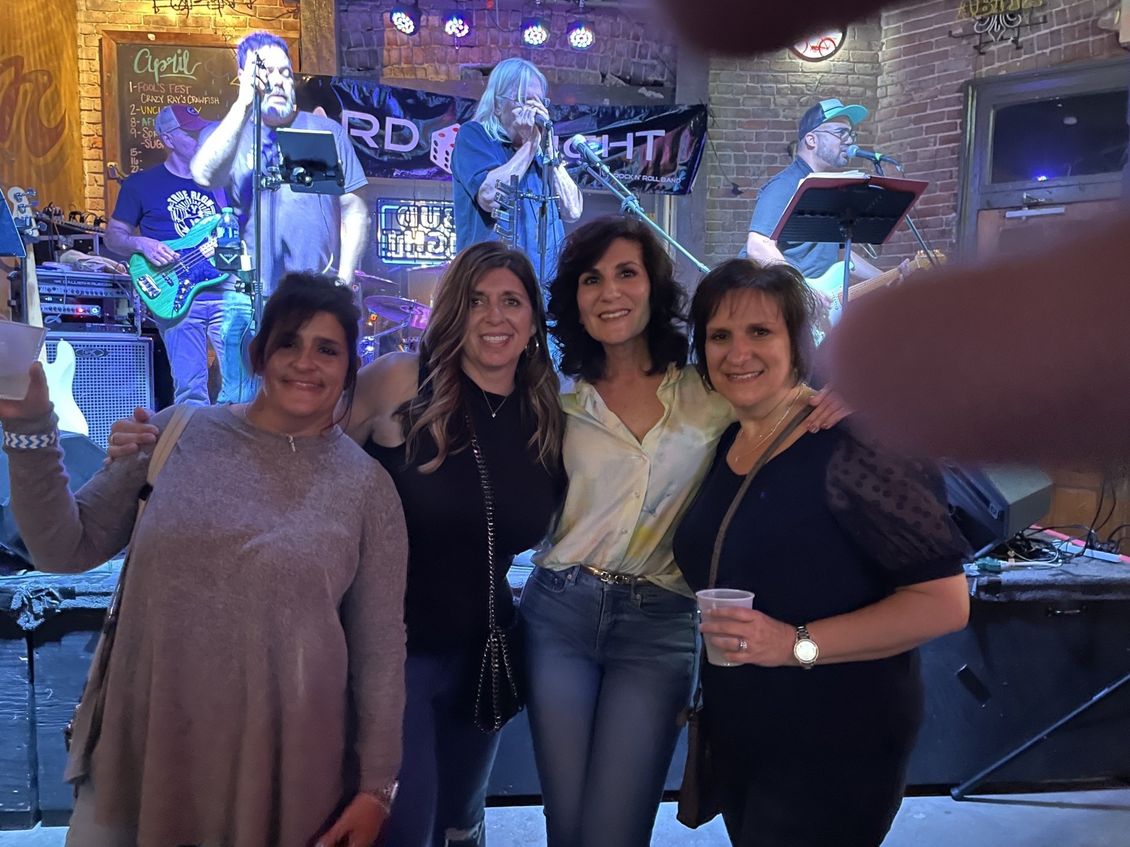 Four women pose in front of a band on stage at a bar.