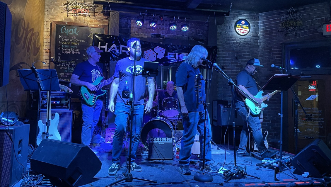 A band of four men performing on a small stage at a bar, lit with blue lights.