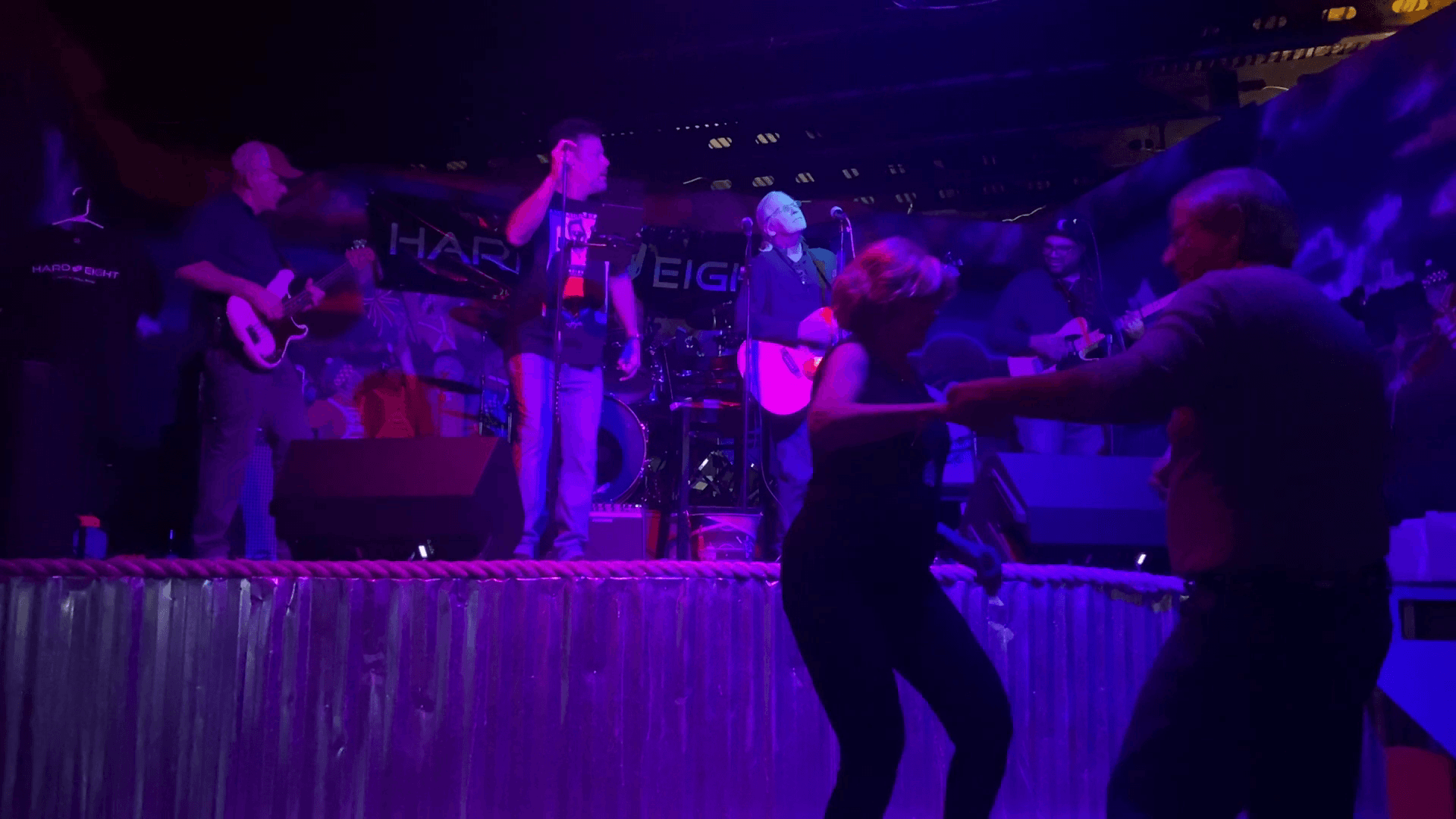 Band performing on stage in a dimly lit club. Couple dances in the foreground, illuminated by blue stage lights.